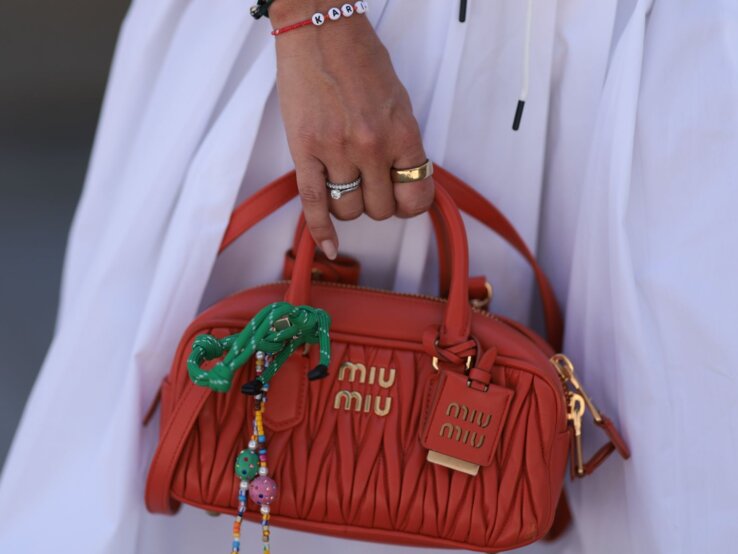 Hand mit goldenen und bunten Armbändern hält eine kleine orangene Miu Miu-Tasche mit dekorativen Elementen, vor einem weißen Kleid.
