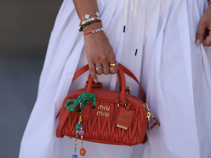Beste Herbst Handtasche Hand mit goldenen und bunten Armbändern hält eine kleine orangene Miu Miu-Tasche mit dekorativen Elementen, vor einem weißen Kleid.
