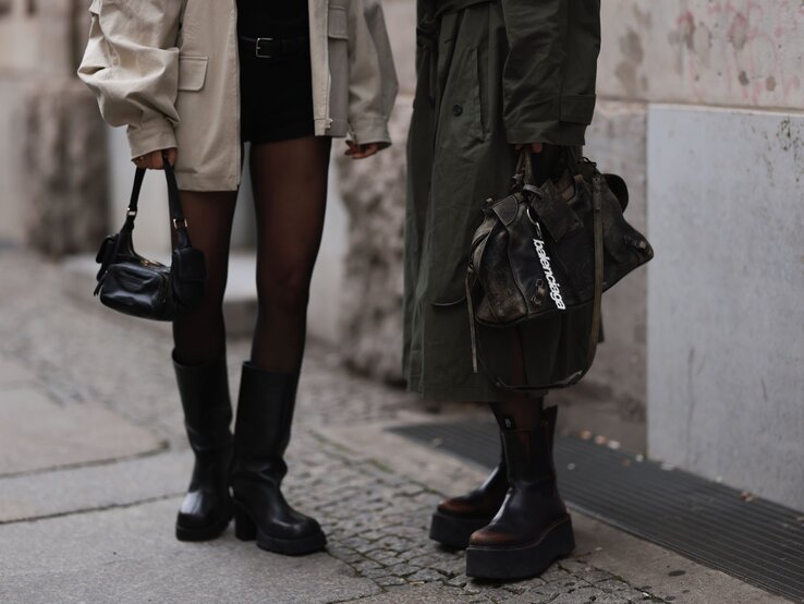 Zwei Frauen stehen auf einer gepflasterten Straße, tragen schwarze Stiefel, kurze und lange Mäntel sowie schwarze Handtaschen.