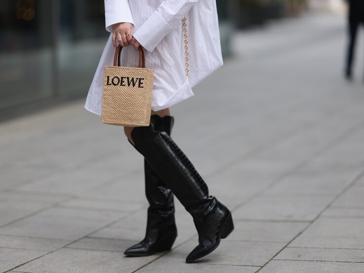 Frau in weißem Hemdkleid mit langen Ärmeln, schwarzen Stiefeln und kleiner geflochtener Tasche von Loewe auf gepflasterter Straße.