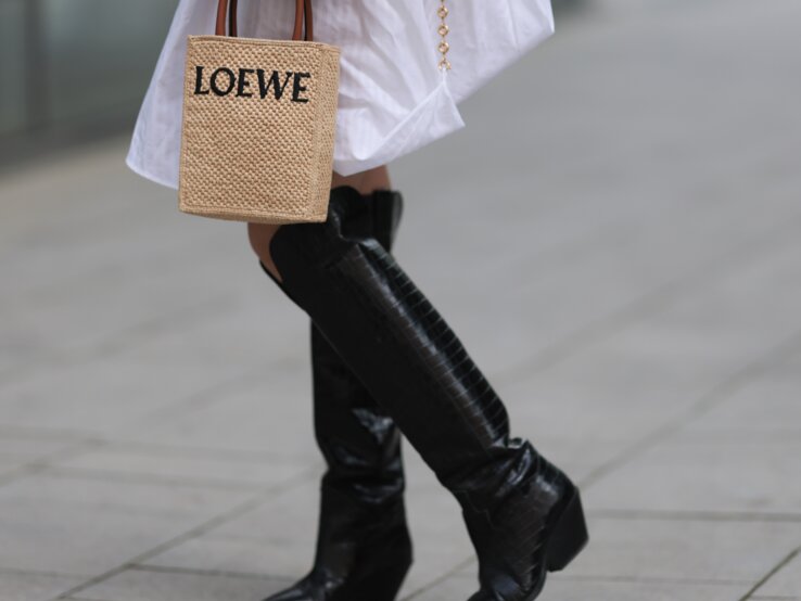 Frau in weißem Hemdkleid mit langen Ärmeln, schwarzen Stiefeln und kleiner geflochtener Tasche von Loewe auf gepflasterter Straße.