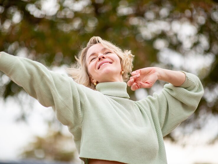 Eine Person mit blondem Haar trägt einen hellgrünen Pullover mit Rollkragen und hebt beide Arme vor einer herbstlichen Baumkulisse.