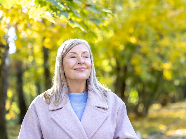 Eine Person mit langen graublonden Haaren trägt einen hellrosa Mantel und einen blauen Pullover im herbstlichen Park.