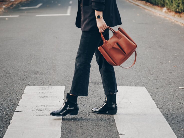Eine Person trägt schwarze Boots, dunkle Jeans und hält eine braune Tasche, während sie einen Zebrastreifen überquert.
