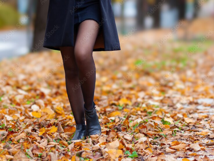 Beine einer Frau in schwarzem Mantel, kurzem Rock und Stiefeletten, gehend über eine von Herbstlaub bedeckte Straße.