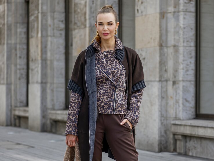 Annika Lau in brauner Hose und Animalprint-Jacke mit passendem Mantel steht vor einer modernen Steinfassade und hält eine flauschige Tasche. | © Streetstyleshooters for QVC