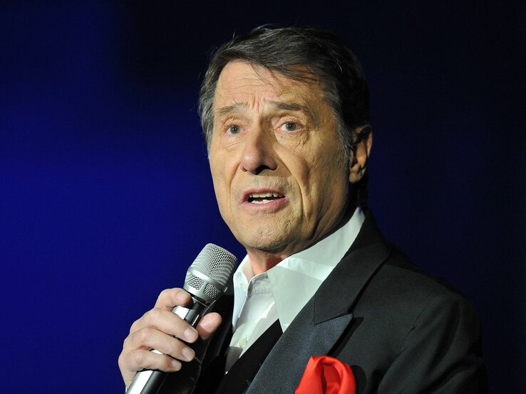 Udo Jürgens trägt einen dunklen Anzug mit rotem Einstecktuch und spricht oder singt in ein Mikrofon vor einem blauen Hintergrund.