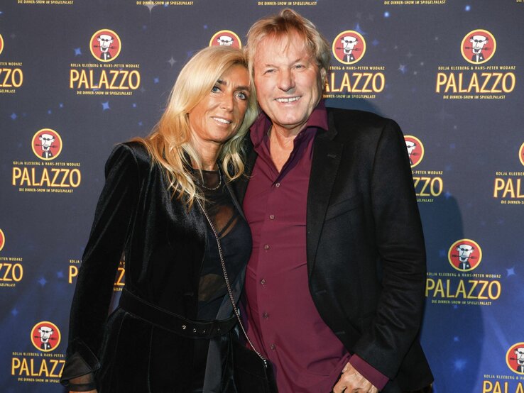 Das Bild zeigt Bernhard Brink und Ute Brink bei einer Veranstaltung auf dem roten Teppich. Hinter ihnen ist ein Hintergrund mit dem Logo "PALAZZO", was darauf hindeutet, dass es sich um eine gesponserte Veranstaltung oder einen Ort handeln könnte, der mit Dinner-Shows oder ähnlichen Unterhaltungsangeboten in Verbindung steht. Sie sind schick gekleidet; Bernhard Brink trägt ein bordeauxrotes Hemd mit einem dunklen Blazer, und Ute Brink trägt eine schwarze Jacke mit passendem Oberteil darunter. Sie scheinen gut gelaunt zu sein und genießen den Anlass.