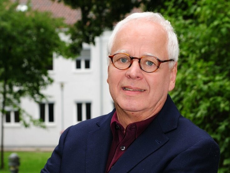 Ein Porträt eines älteren Herren mit grauem Haar und runden Brillen. Er trägt ein dunkelblaues Sakko über einem burgunderroten Hemd. Der Mann blickt direkt in die Kamera und lächelt leicht. Im Hintergrund ist ein unscharfes grünes Laubwerk zu sehen, was auf eine Aufnahme im Freien schließen lässt, mit einem weißen Gebäude im Hintergrund.