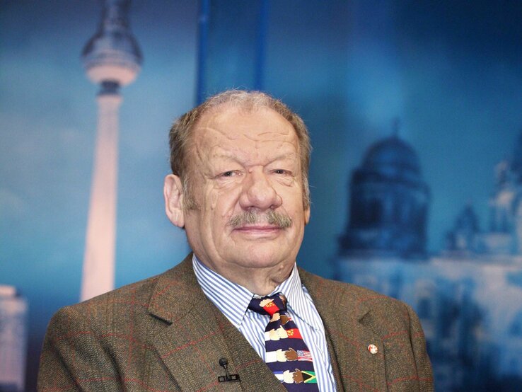 Schauspieler Wolfgang Völz im Anzug schaut ernst in die Kamera, im Hintergrund sind Reichstag und Berliner Fernsehturm als Kulisse zu sehen