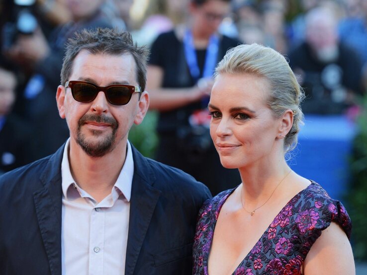 Alex Silva und Nina Hoss stehen Seite an Seite und blicken in die Kamera. Alex Silva trägt eine schwarze Jacke und Sonnenbrille, während Nina Hoss ein Kleid mit Blumenmuster und ein dezentes Make-up trägt. Ihr Haar ist zu einem eleganten Knoten gesteckt. Sie scheinen auf einem roten Teppich zu stehen, erkennbar an den unscharfen Gestalten und Kameras im Hintergrund, was auf eine Film- oder Unterhaltungsveranstaltung hindeutet.