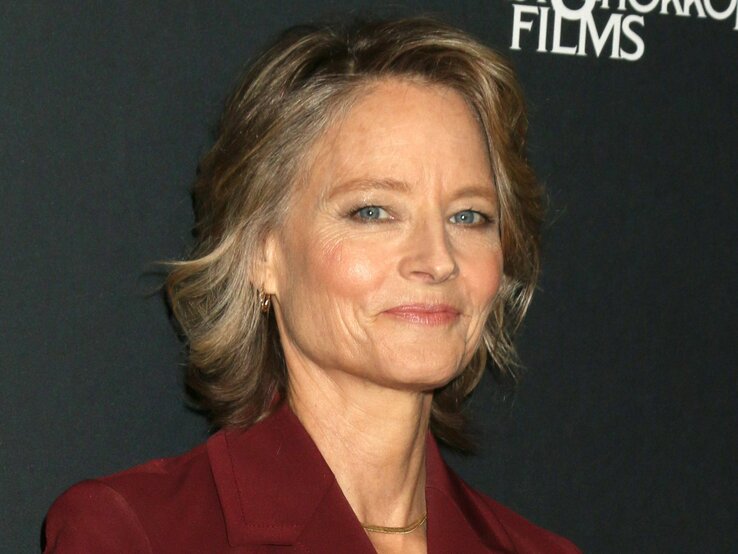 Jodie Foster hat mittellanges graues Haar und trägt einen burgunderfarbenen Blazer. Sie lächelt sanft, aufgenommen vor einem dunklen Hintergrund.