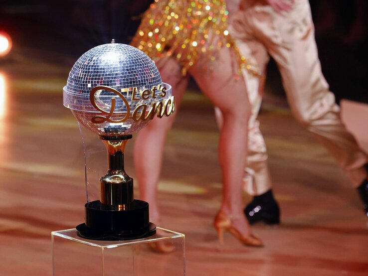 Ein glitzernder Disco-Ball-Trophäe auf einem Podest mit der Aufschrift "Let's Dance", im Hintergrund unscharf die Beine zweier Tänzer in Bewegung, einer trägt schwarze Schuhe und der andere goldene Tanzschuhe mit Glitzer.