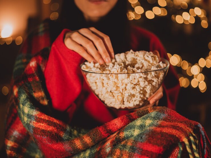 Weihnachtsfilme Redaktion Eine Person in rotem Pullover und karierter Decke greift in eine Schüssel Popcorn vor unscharfen, warm leuchtenden Lichterketten, während sie einen Weihnachtsfilm schaut.