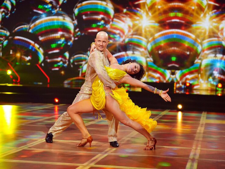 Fabian Hambüchen in einem goldenen Anzug hält bei Let's Dance seine Tanzpartnerin Anastasia Maruster in einem gelben, fransigen Kleid, während sie sich in einer dramatischen Pose zum Tanz neigen.