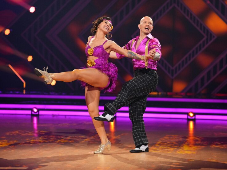 Anastasia Maruster mit goldenen Blüten am Kleid hebt das Bein, Fabian Hambüchen führt sie dynamisch über das bunte Tanzparkett.