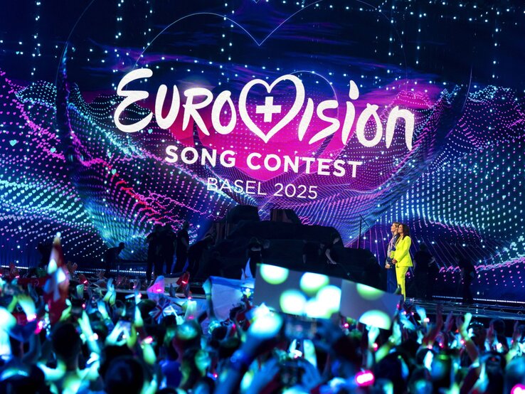 Blick auf die bunte Bühne des Eurovision Song Contest 2025 in Basel mit jubelndem Publikum und zwei Moderatorinnen rechts.