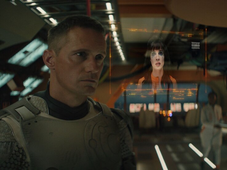 Murderbot Alexander Skarsgård Interview | © Steve Wilkie/ Apple TV+ Alexander Skarsgård in einem futuristischen Raumanzug sieht nachdenklich auf ein holografisches Bild einer Frau mit markanten Make-up, das vor ihm schwebt. | © Steve Wilkie/ Apple TV+
