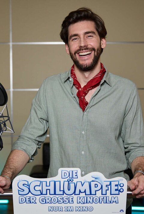 Alvaro Soler mit braunem Bart und grünem Hemd steht vor Mikrofon, während er für "Die Schlümpfe" Werbung macht.