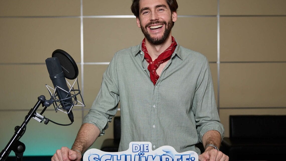 Alvaro Soler mit braunem Bart und grünem Hemd steht vor Mikrofon, während er für "Die Schlümpfe" Werbung macht.