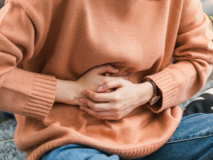  Das Bild zeigt den Mittelteil einer Person, die einen pfirsichfarbenen Strickpullover und eine Uhr trägt und sich mit gefalteten Händen auf dem Bauch hält, was möglicherweise auf Bauchschmerzen oder Unbehagen hinweist.