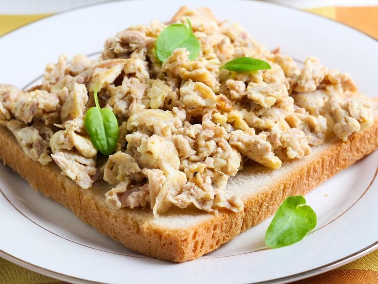 Ein belegtes Toastbrot mit Thunfisch-Rührei garniert mit frischen grünen Blättern, wahrscheinlich Feldsalat, auf einem weißen Teller mit einem goldenen Rand, serviert auf einer gelb gemusterten Tischdecke.