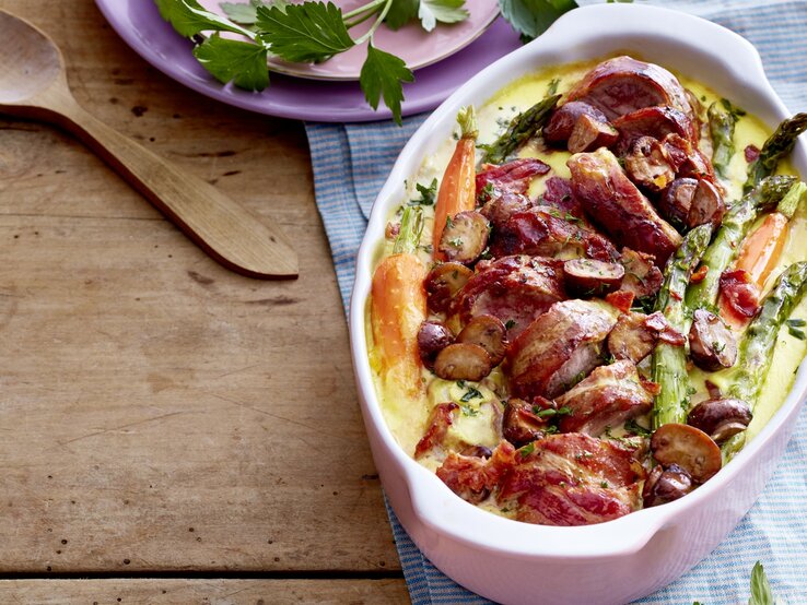 Schweinefilet mit Frühlingsgemüse in einer weißen Auflaufform, daneben Teller mit Petersilie und Holzlöffel auf Holzuntergrund. 