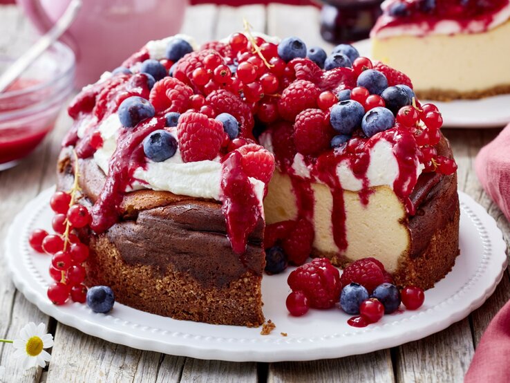 Ein angeschnittener Beeren-Käsekuchen mit Schlagsahne und verschiedenen frischen Beeren darauf auf einem weißen Teller und heller, verschwommener Hintergrund. 