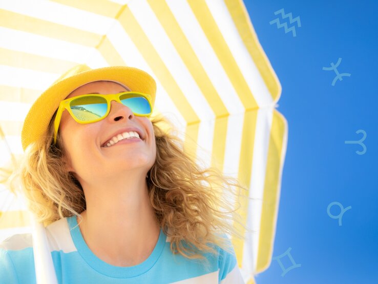 Das Bild zeigt eine lächelnde Frau mit lockigen blonden Haaren, die eine gelbe Sonnenhut und gelbe Sonnenbrille trägt. Sie steht unter einem gelb-weiß gestreiften Sonnenschirm vor einem klaren, blauen Himmel. Auf dem Himmel sind leicht transparente Symbole verschiedener Sternzeichen zu sehen. Die Stimmung ist sommerlich und fröhlich.