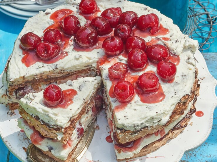Angeschnittene Schwarzwälder Kirsch-Baisertorte mit Kirschen oben drauf auf blauem Untergrund. 