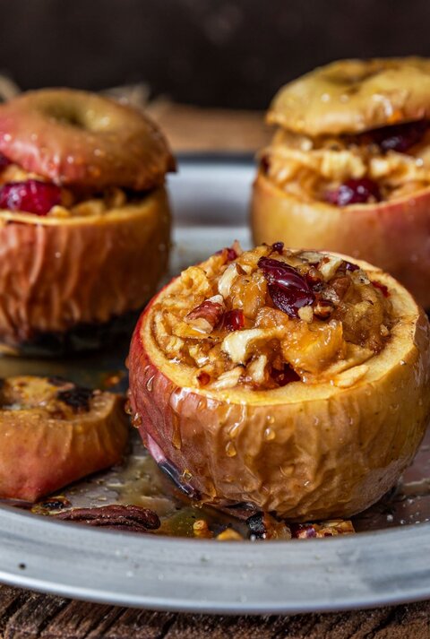 Drei ofengebackene Äpfel mit nussiger Füllung und Cranberries in einem Metallteller, dekoriert mit Zimtstangen auf rustikalem Untergrund.