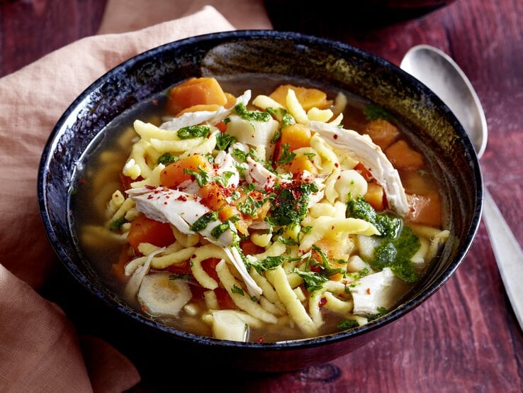 Dieser Hühnertopf mit Spätzle bringt alles mit: Saftiges Fleisch, frisches Gemüse und Spätzle, die jede Schale in ein Highlight verwandeln. Lass es dir schmecken! Hühnertopf mit Spätzle in einer dunkelblauen Schüssel auf hölzernem Hintergrund. Links ein rosa Geschirrtuch, rechts ein Löffel.