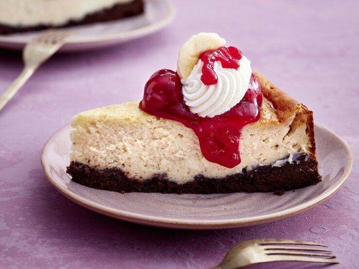Ein Stück KiBa-Cheesecake auf einem Teller auf rosa Hintergrund. Vorne auf dem Foto eine Gabel.