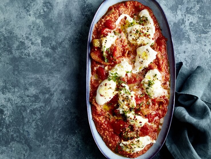 Zerzupfter Büffelmozzarella auf Röstgemüse à la Romesco in einer Auflaufform auf dunklem Hintergrund.