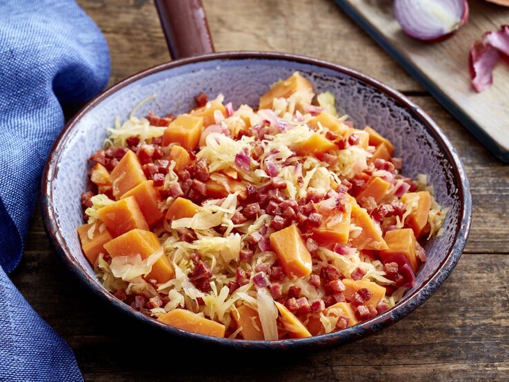 Knuspriger Schinken, zarte Süßkartoffel, würziges Sauerkraut – diese Süßkartoffel-Sauerkraut-Pfanne hat alles, was du brauchst! Süßkartoffel-Sauerkraut-Pfanne auf hölzernem Hintergrund. Links davon ein blaues Geschirrtuch.