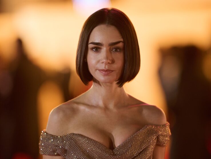Lily Collins mit kurzen dunklem Blunt Bob und funkelndem, goldenen Abendkleid steht ernst vor warm beleuchtetem Hintergrund.