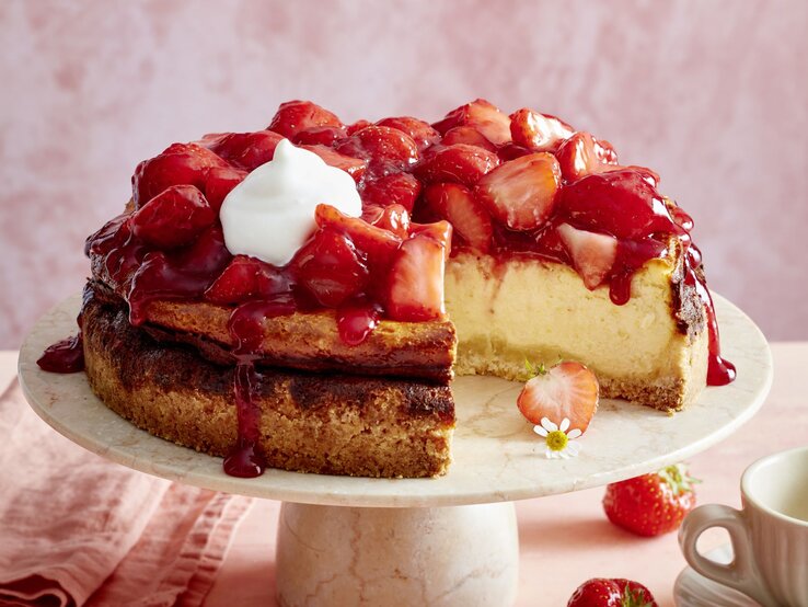 Erdbeer-Käsekuchen mit Sahne auf einem beigen Tortenteller vor rosa Hintergrund, darauf Erdbeeren und eine Tasse Kaffee verteilt. 