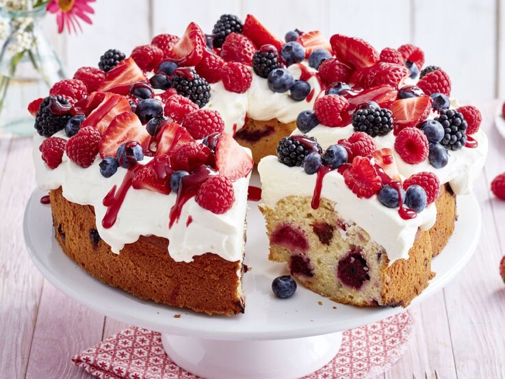 Beeren-Schmand-Kranz auf einem weißen Tortenteller auf einem rot-weißen Tuch vor hellem Hintergrund. 