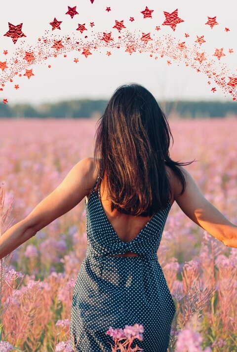 Eine Frau mit dunkelbraunem Haar in einem ärmellosen, gepunkteten Kleid steht mit dem Rücken zur Kamera in einem weiten Feld voller rosa-lila Wildblumen. Ihre Arme sind seitlich ausgestreckt, während sie die Blumen berührt. Um sie herum ist ein digital eingefügter Halbkreis aus roten Sternen zu sehen, der wie ein magischer Funkenbogen wirkt. Der Himmel ist hell und klar, im Hintergrund ist eine Baumlinie zu erkennen.