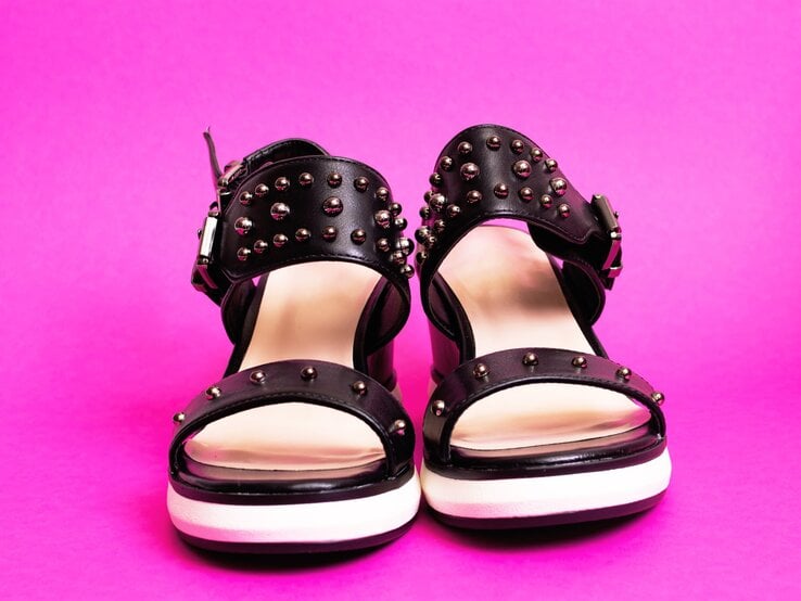 Sandalen-DIY Schwarze Sandalen mit silbernen Nieten und weißen Sohlen stehen frontal vor pinkem Hintergrund.