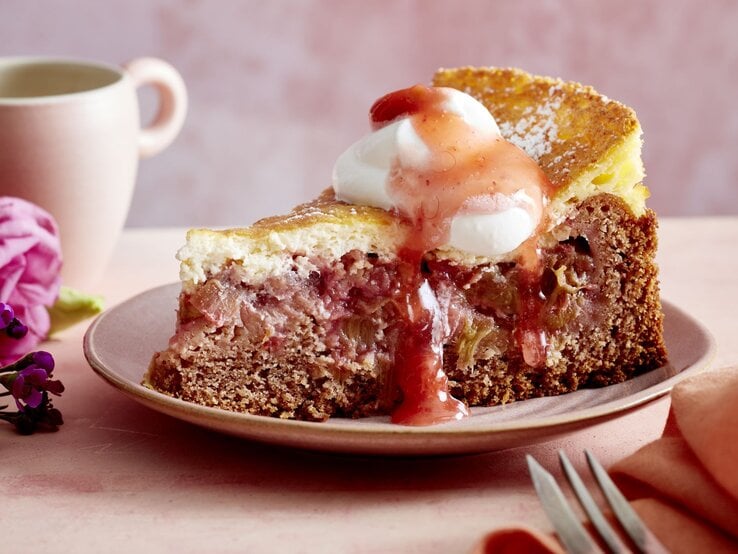 Mit dieser Torte bekommt Rhabarber ein cremiges Update – locker gebacken, frisch im Geschmack und perfekt für Frühling und Sommer. Probiere jetzt die Rhabarber-Torte mit Schmand! Ein Stück Rhabarber-Torte mit Schmand auf einem rosa Teller vor rosa Hintergrund. Hinten auf dem Foto eine Tasse.