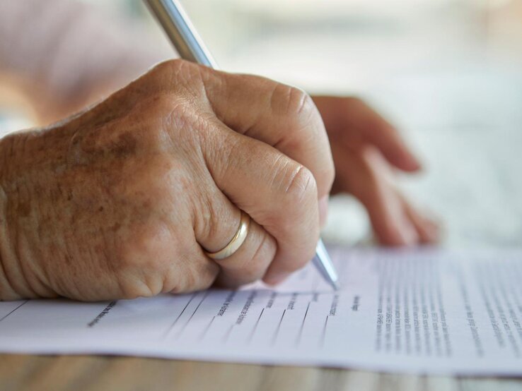 Nahaufnahme einer Hand einer älteren Dame, die mit einem Stift ein Formular ausfüllt. 