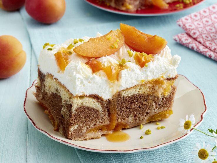 Kuchen fürs Herz: Diese Aprikosen-Marmor-Schnitten machen glücklich (versprochen) Ein Stück Aprikosen-Marmor-Schnitten auf einem Teller: saftiger Marmor-Rührteig mit Aprikosen, luftiger Sahnehaube, karamellisierten Fruchtstücken und gehackten Pistazien als Topping. Im Hintergrund frische Aprikosen auf hellblauem Untergrund.