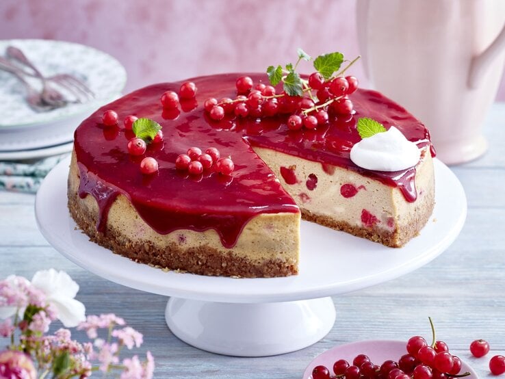  Johannisbeer-Cheesecake auf einem weißen Tortenteller vor hellem Hintergrund. Hinten auf dem Foto kleine Teller, vorne Blumen und Johannisbeeren.