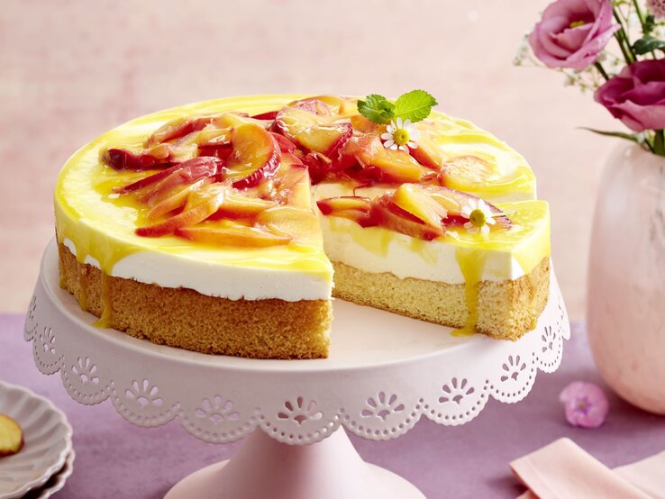 Cremiger Joghurt, saftige Nektarinen und ein luftiger Boden – die Nektarinen-Joghurt-Torte ist leicht zuzubereiten und angenehm erfrischend. Nektarinen-Joghurt-Torte auf einem weißen Tortenteller vor rosa und lila Hintergrund. Links kleine Teller und rechts auf dem Foto Blumen in einer Vase.