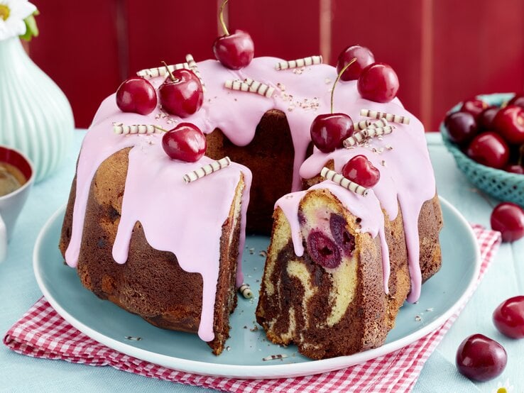Ein saftiger Kirsch-Marmor-Hupf mit rosa Zuckerguss, frischen Kirschen und Schokoröllchen dekoriert, auf einem hellblauen Teller und rotem Untersetzer serviert. Im Anschnitt sind die Marmorierung und eingebackene Kirschen deutlich zu sehen.
