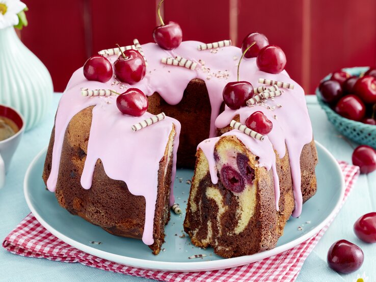 Mit Kirsch-Marmor-Hupf wird jeder Tag zum Kuchentag – gönn dir den süßen Twist mit Kirsch und Kakao! Ein saftiger Kirsch-Marmor-Hupf mit rosa Zuckerguss, frischen Kirschen und Schokoröllchen dekoriert, auf einem hellblauen Teller und rotem Untersetzer serviert. Im Anschnitt sind die Marmorierung und eingebackene Kirschen deutlich zu sehen.