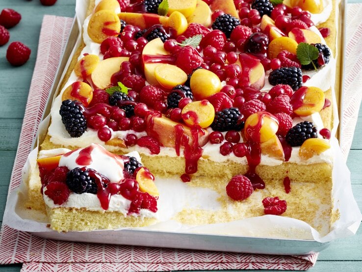 Tutti Frutti Schnitten auf einem weißen Blech auf einem rot-weißen-Geschirrtuch vor grünem Hintergrund. Hinten auf dem Foto Himbeeren.