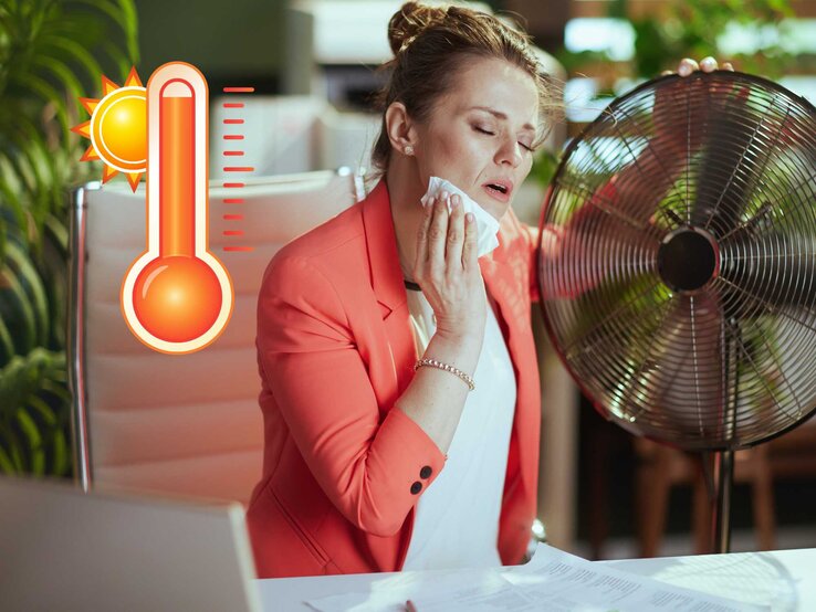 Frau sitzt im Büro an einem Ventilator, weil ihr heiß ist. 
