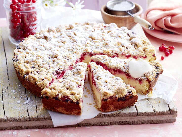 Johannisbeer-Streusel-Käsekuchen Johannisbeer-Käsekuchen mit Streuseln auf einem Holzbrett serviert, ein Stück angeschnitten, daneben ein Glas mit roten Johannisbeeren und Gänseblümchen.